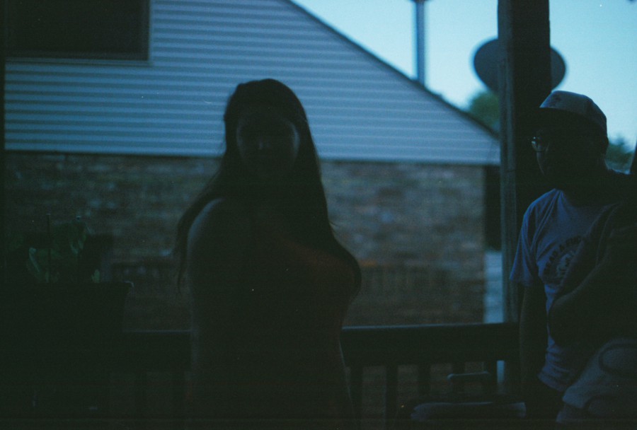 A silhouetted figure, Ryanne, is standing on a back porch, her form faintly visible against the dim lighting of a dusky sky. A man is to her right, partially visible, wearing a baseball cap. The background includes a slanted rooftop and a satellite dish, all against the faint blue of a Chicago neighborhood evening.