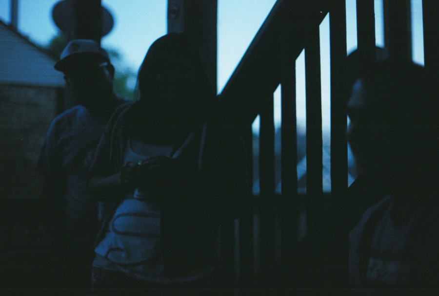 A group of three people standing on a back porch, their silhouettes outlined by fading evening light. The porch railing and vertical beams of the structure frame the composition. The people are difficult to distinguish due to the film’s low-light grain, adding an atmospheric effect to the scene.