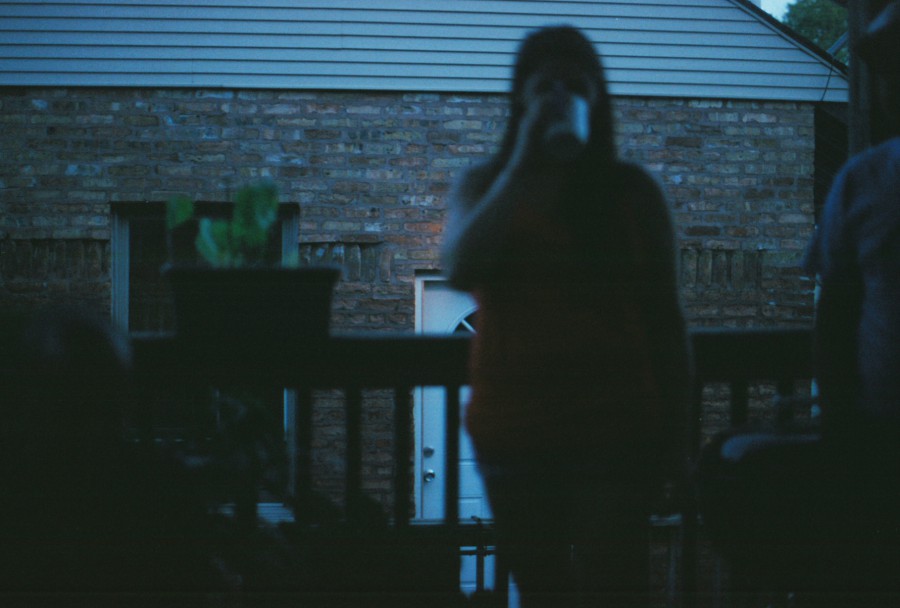 Ryanne stands on the back porch with her figure slightly in shadow, sipping from a cup. A houseplant in a pot sits on the porch railing, and the background features the sharp details of a red-brick building, with a white door visible below, giving the image a layered perspective.
