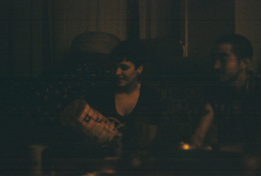 Indoors, Emily and Alex are seated on a couch in a dimly lit room. Emily holds an object and smiles, her short dark hair framing her face. Alex sits to her right, faintly visible. The room is warmly lit but dark, adding a moody and intimate feel captured on 35mm film.