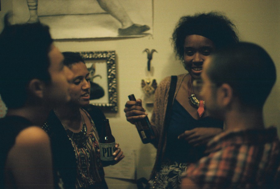 Four people stand in a lively discussion during the party, their faces warmly illuminated. A woman in a blue dress with a bold necklace smiles as she holds a beer, while another person gestures with a bottle. The room has eclectic décor, including a framed piece of art and a decorative hook, adding character to the setting.