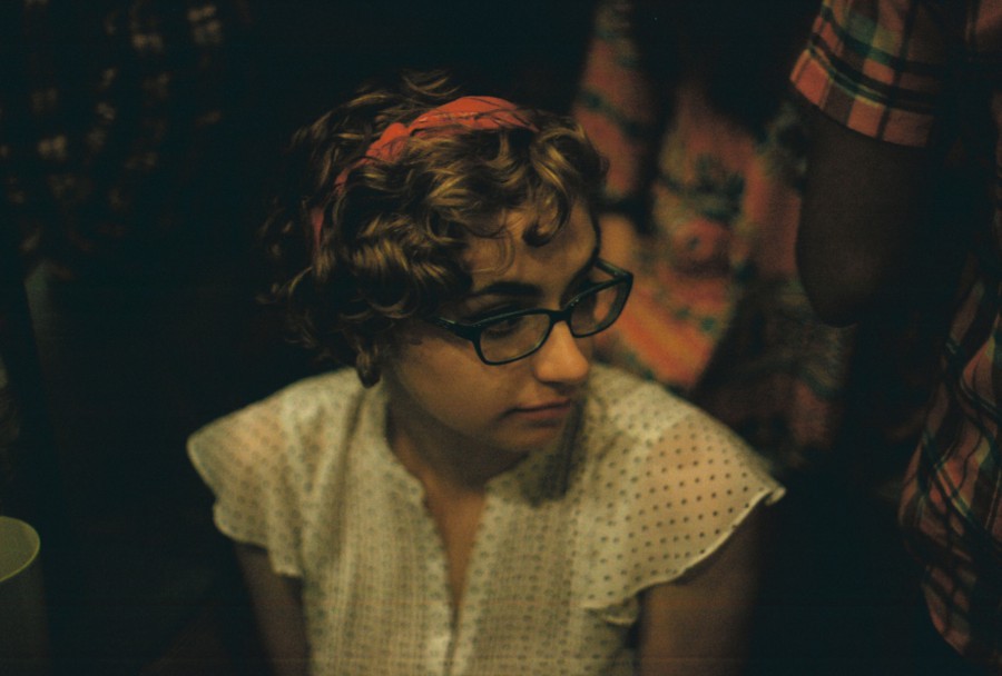 A candid photo of Katie at the party, captured from a slightly higher angle. She has short, curly hair with a red headband, wearing glasses and a vintage-style white blouse with small polka dots. The background features muted, colorful clothing patterns of other attendees, slightly out of focus due to the shallow depth of field provided by the 35mm film SLR camera.