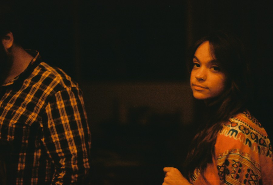 A warm-toned nighttime photo featuring a man on the left in a yellow and black plaid shirt, his face partially turned away, and Caitlin on the right in a patterned, brightly colored top. Caitlin looks directly at the camera with a subtle, knowing expression, her long, dark hair framing her face. The dimly lit background is dark and indistinct.