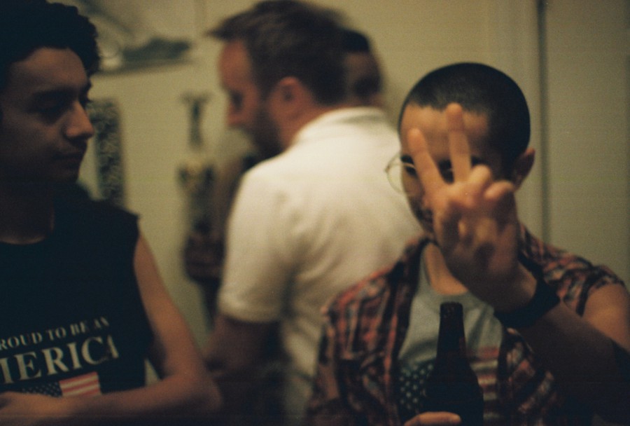 Alex’s brother on the left, wearing a black sleeveless shirt with white text that reads, "Proud to be an American," stands in conversation with Alex, who is on the right, holding up a peace sign with his right hand while gripping a beer bottle in the other. Behind them, several people mingle in a warmly lit indoor space. The background includes subtle wall decorations, creating a casual party atmosphere.