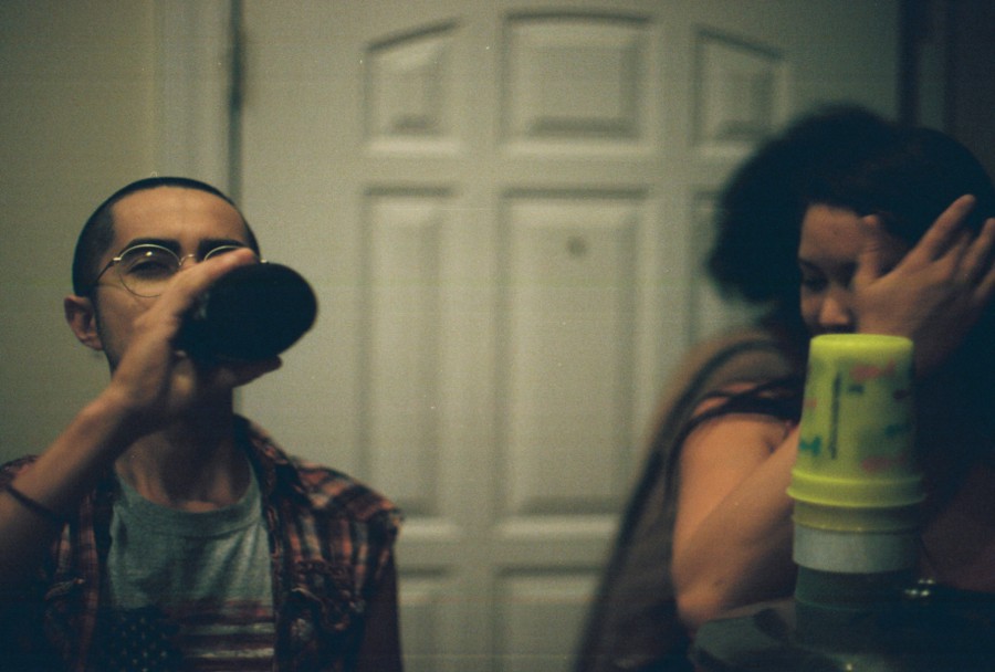 Alex, wearing a plaid shirt and round glasses, drinks from a dark beer bottle while standing in front of a white door. Ryanne is partially visible on the right, covering her face with her hand, while another person is slightly blurred in the background. The dim, warm lighting adds a cozy yet intimate ambiance.