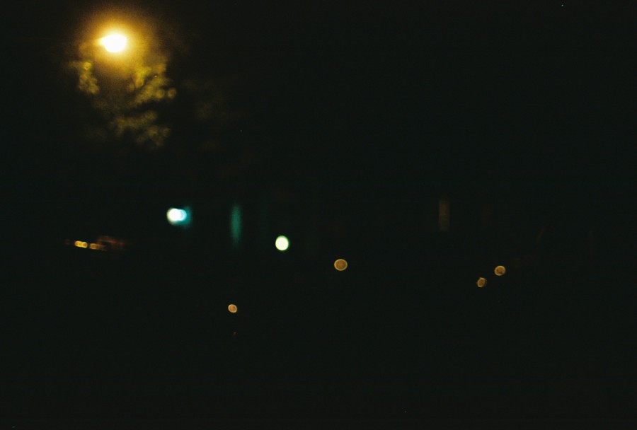A dimly lit city street at night, with glowing orbs of streetlights and house lights punctuating the darkness. The soft, blurred effect captures an almost abstract representation of urban life after dark, emphasizing the warm glow of the lights.