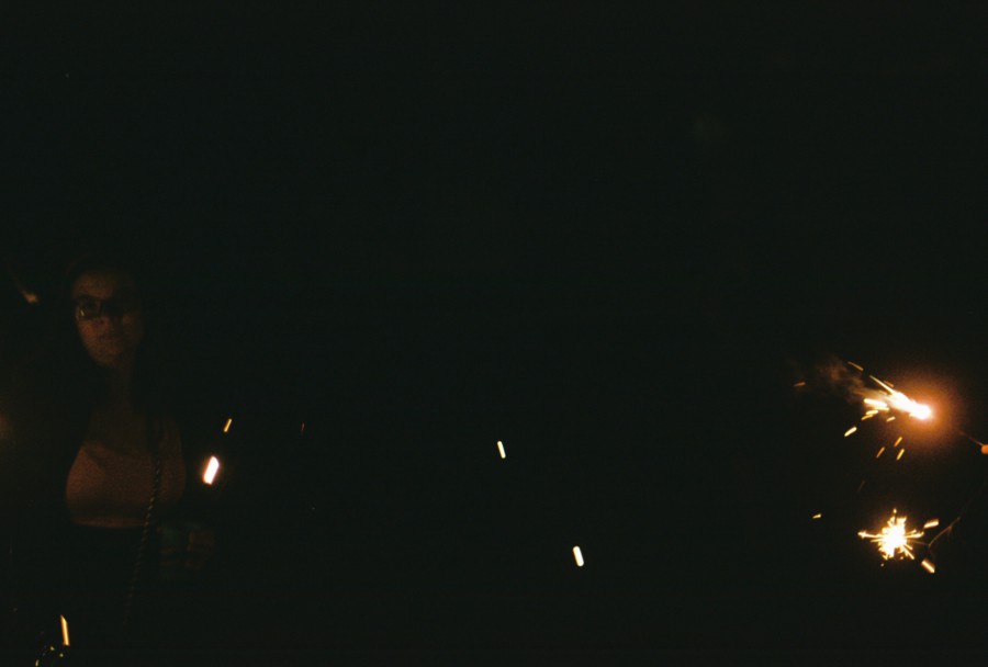 A street scene at night with a person faintly visible in the darkness. Bright fireworks sparkle to the right, scattering orange and white sparks into the air. The sparse lighting and silhouettes create a mysterious, celebratory mood.