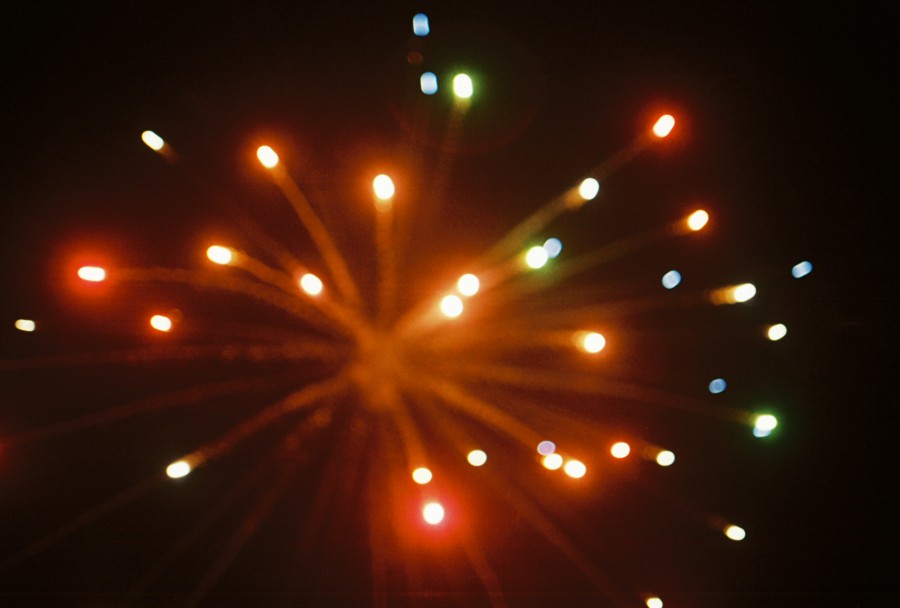 A vibrant, dynamic shot of a firework bursting in the night sky. Bright streaks of red, green, orange, and blue light radiate outward, creating a mesmerizing, blurred effect as the colorful explosion dominates the dark background. The soft focus and 35mm film grain enhance the sense of energy and celebration.