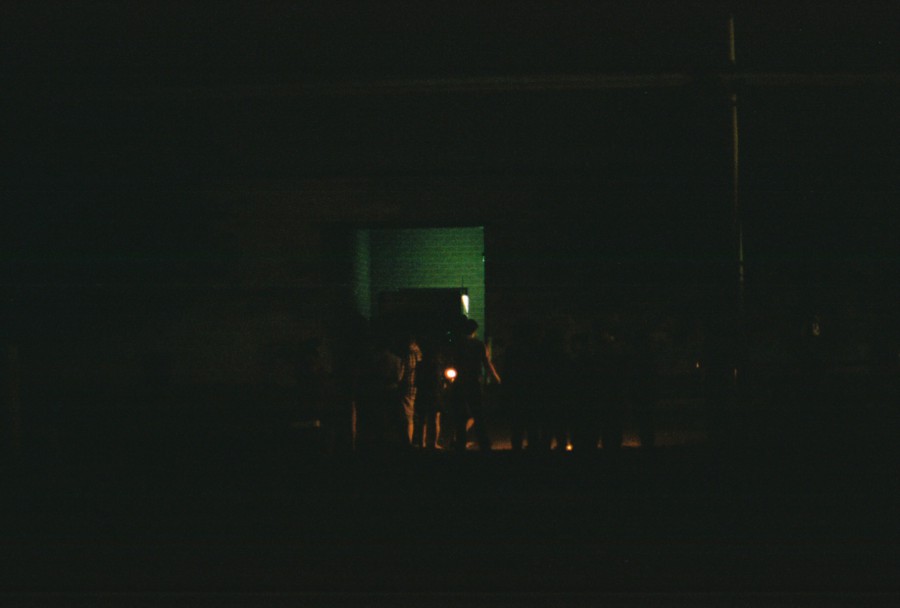 A dark street scene showing a group of people gathered near a lit doorway. The doorway emits a greenish light that slightly illuminates the figures standing and conversing outside. The surrounding area is dimly lit, with shadows obscuring most details. The silhouette of individuals suggests casual summer attire, reflecting a warm evening in Chicago West Town during the Fourth of July celebration. Shot on 35mm film with an SLR camera, the grain adds a nostalgic, atmospheric quality to the image.