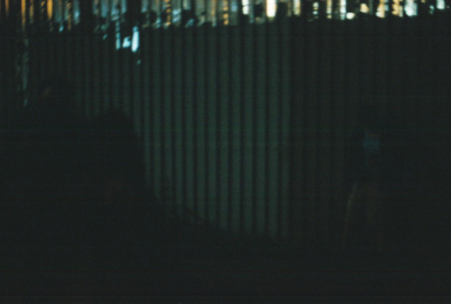 A metal ribbed wall stretches across the frame in a dark environment, with faint lighting creating shadows and highlights on the surface. On the right side, a barely discernible figure stands in partial shadow, wearing what appears to be a light top and darker pants. The upper part of the image reveals hints of industrial structures and dim city lighting. The photograph is moody and textured, captured with a 35mm SLR camera, emphasizing the interplay between light and darkness.