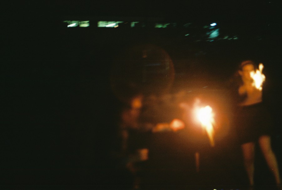 A blurred shot of two people holding sparklers at night. The sparklers create bright, dynamic trails of light that dominate the frame, casting warm orange and yellow tones. The individuals are faintly visible, with one on the right wearing a dark skirt and the other slightly obscured in motion. A faint halo effect around the sparklers adds to the dreamy, celebratory mood of the Fourth of July. The film’s grain enhances the warm, analog aesthetic.