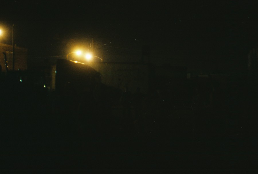 A minimal nighttime cityscape with deep darkness enveloping most of the frame. A couple of bright streetlights punctuate the scene, their glow illuminating vague outlines of buildings and distant structures. The lights create a stark contrast against the blackness, emphasizing the quiet and stillness of the urban night. The 35mm film grain adds subtle texture, enhancing the moody and subdued atmosphere of this Chicago West Town evening.