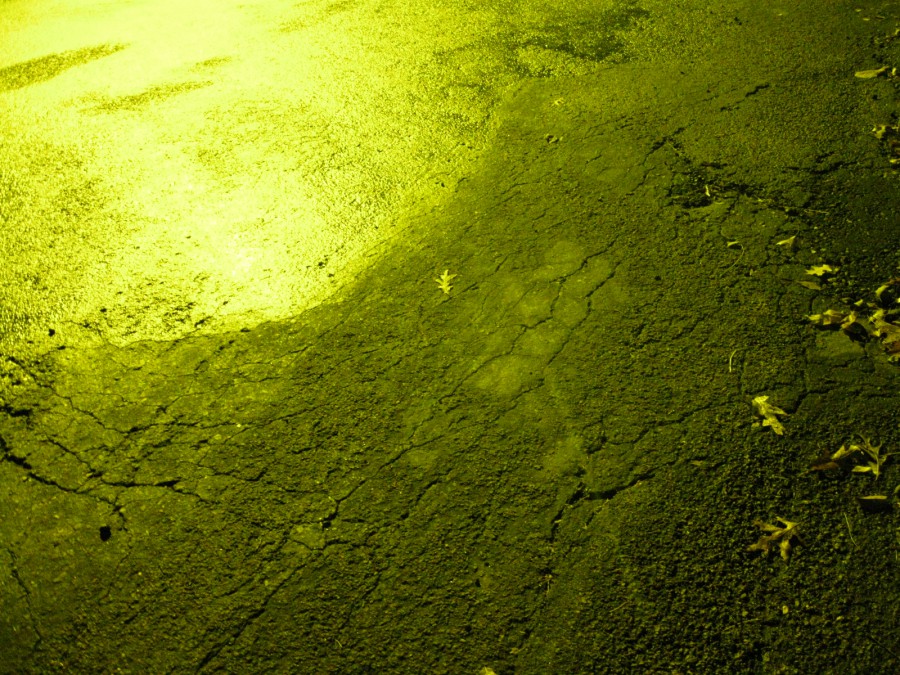 A closer image of cracked asphalt, illuminated in yellow-green tones from a nearby streetlight. The texture of the road surface is rough, with small leaves scattered across the picture. Digital photograph taken on a Canon PowerShot A1000 IS in northeast Eau Claire, WI, in the Spring of 2011.