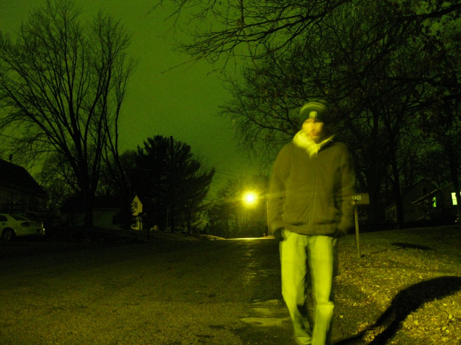 Me, Ethan Hulbert, in a green stocking cap, brown jacket, and blue jeans, slightly blurred to imply motion, walking down a residential street, illuminated by one single yellow street lamp in the background. Silhouettes of tall bare trees, homes, the bare road, leaves on the sidewalk, and some ice and snow left on the road surround me. Digital photograph taken on a Canon PowerShot A1000 IS in northeast Eau Claire, WI, in the Spring of 2011.