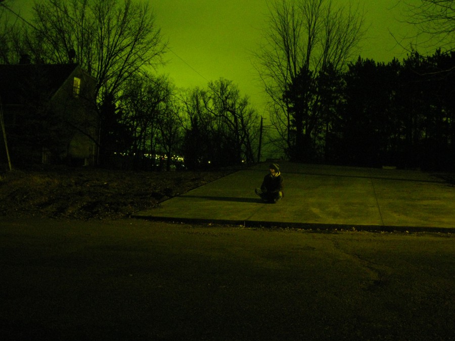 A residential street at night, along with a cement or asphalt flat surface where I sit, a young man in a brown jacket and beanie, small in the overall photo, framed by bare tree branches and a greenish sky. My shadow is cast long on the cement. Digital photograph taken on a Canon PowerShot A1000 IS in northeast Eau Claire, WI, in the Spring of 2011.