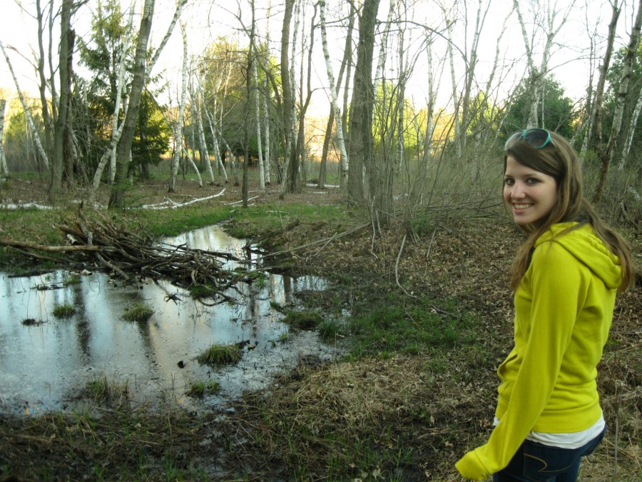 A beautiful reflective pond in the woods, surrounded by tall birch trees and lots of green and brown low undergrowth. A young woman - Katie Rodgers (or "krod") - stands in the foreground facing the pond, but turning back towards the camera with a bright smile. She wears a bright yellow hoodie and blue sunglasses on her long brown hair. The overall mood is cheerful and serene. Digital photograph taken on a Canon PowerShot A1000 IS in northeast Eau Claire, WI, in the Spring of 2011.