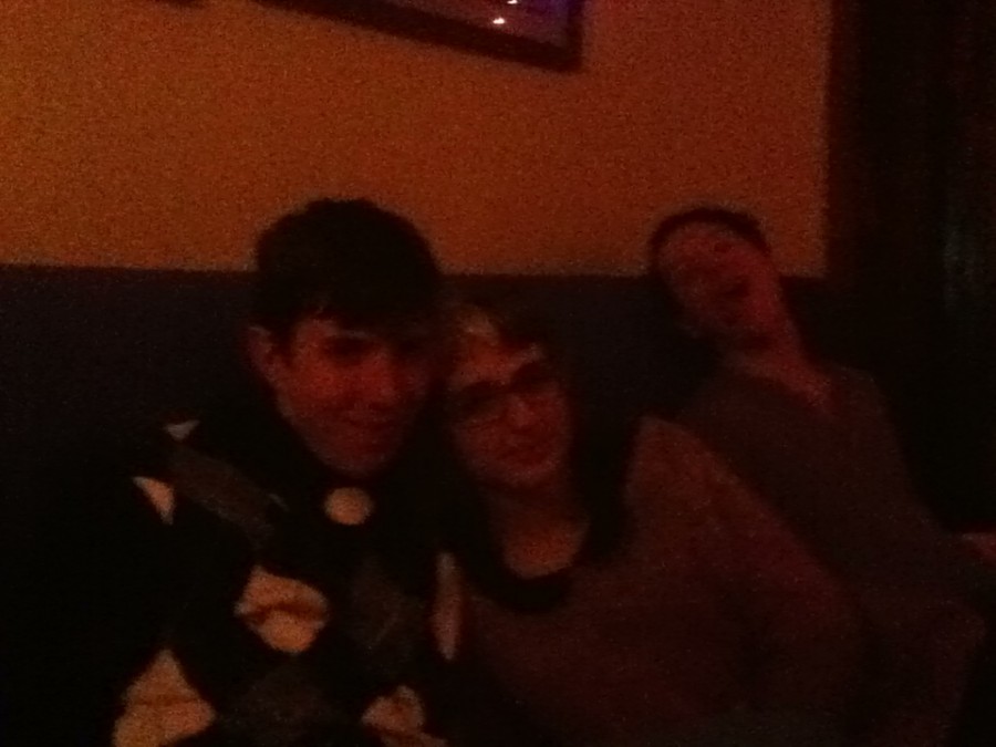 A candid group shot taken in low lighting, showing three people sitting on a blue couch. On the left, a man named Bob wears a dark argyle sweater, his arm around Lauren, who has distinctive split-dyed hair—half blonde, half dark—and wears glasses and a cozy sweater. They sit close together, appearing affectionate and comfortable. In the background, Brandon leans back with an exaggerated, wide-mouthed laugh, adding a playful and casual element to the scene. The image is dark and grainy, with warm, low-quality lighting typical of a bar or lounge setting.