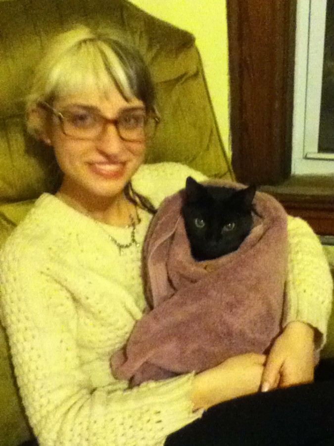 Lauren sits in a large, vintage armchair, holding Callie, a sleek black cat, swaddled in a soft, faded purple towel. Lauren has split-dyed hair (half blonde, half dark brown) and wears glasses along with a cream-colored knit sweater. She smiles warmly at the camera, exuding a relaxed and happy vibe. The background features a wood-paneled window with soft, warm lighting, emphasizing the cozy, homely atmosphere. The image has a slightly yellowish tint, common with indoor lighting and older phone cameras.