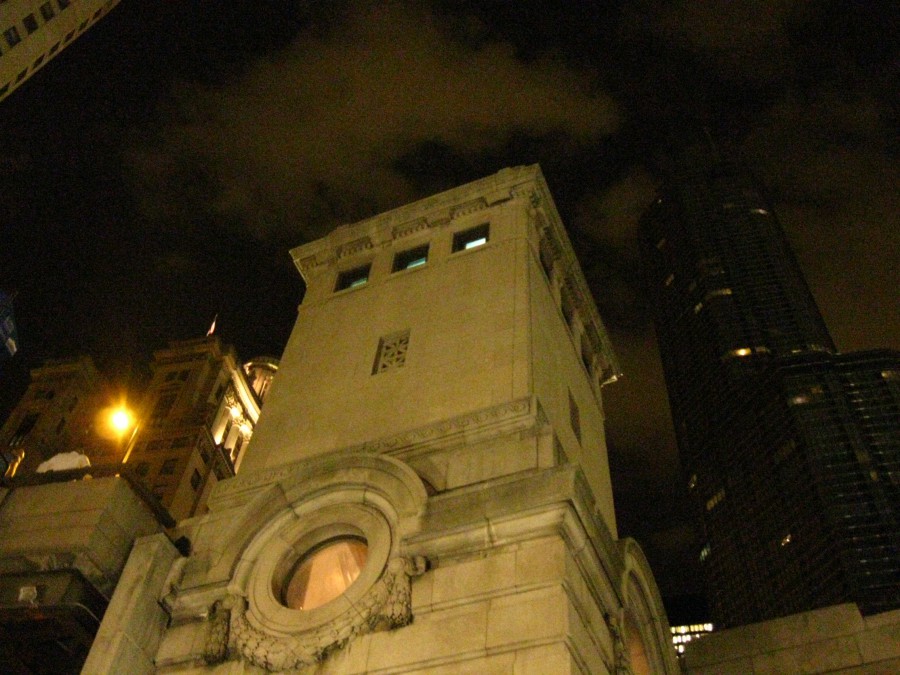 A low-angle view of a Chicago river bridge structure, focusing on a large, ornate architectural guard tower with rectangular windows near the top. Surrounding it, taller modern skyscrapers loom in the background, with a cloudy night sky above. The scene blends historical architecture with urban modernity, framed by a dim yet dramatic light.