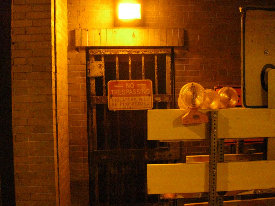 A barred doorway lit by a bright yellow-orange light above, casting harsh shadows on the surrounding brick wall. A prominent red "No Trespassing Violators Will Be Prosecuted" sign hangs on the gate, while construction-style barricades with reflective lights sit in front. The image emphasizes restriction and caution, matching the night's ghost-hunting theme.