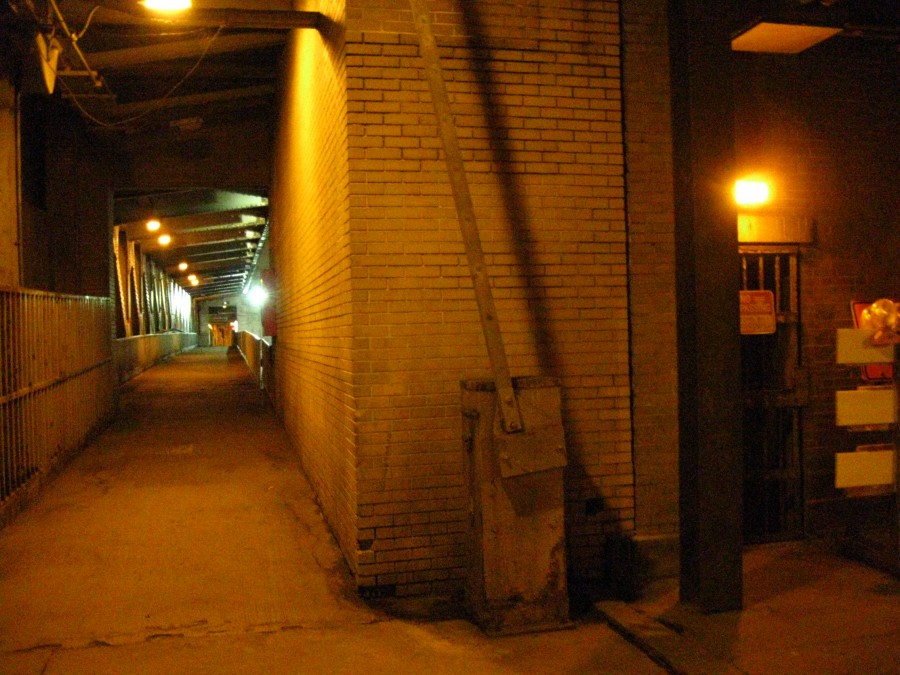 A long tunnel illuminated by a series of yellow-orange lights, creating a moody, dimly lit urban atmosphere. The left side features a metal railing extending into the distance, while the right side showcases a flat brick wall with a small gated area and a metal utility box. Emergency barriers and a barred gate with no trespassing signs are visible in the foreground. The perspective emphasizes the tunnel's depth and infrastructure details.