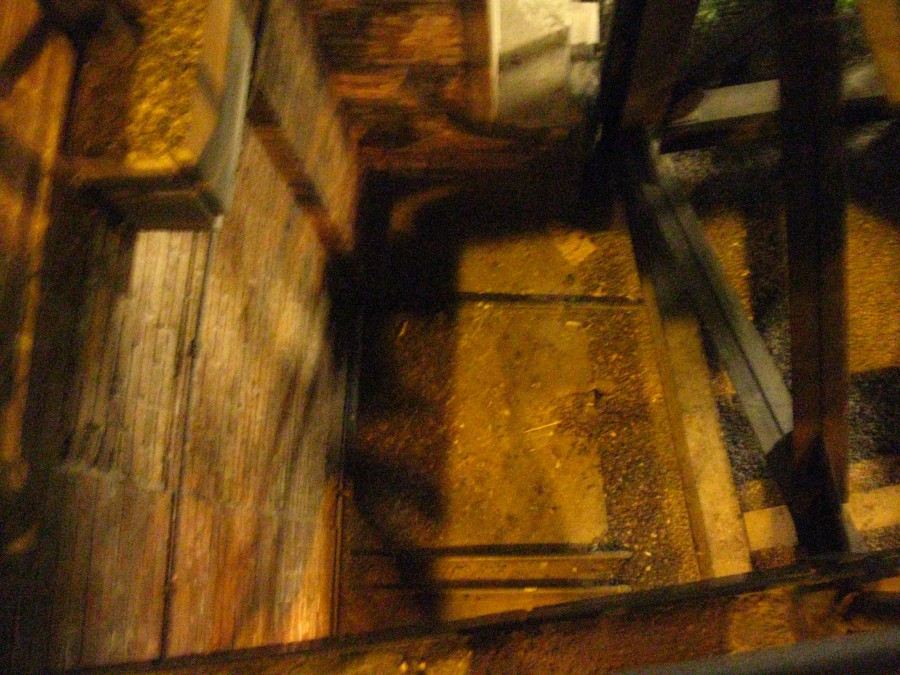 A blurry, motion-heavy image looking downward into a pit-like structure. Wooden beams and metal supports frame the edges of the shot, while the center shows a dirt and gravel surface. The photo's lack of sharp focus and dim lighting create an unsettling, ambiguous effect, contributing to the ghost-hunting theme.