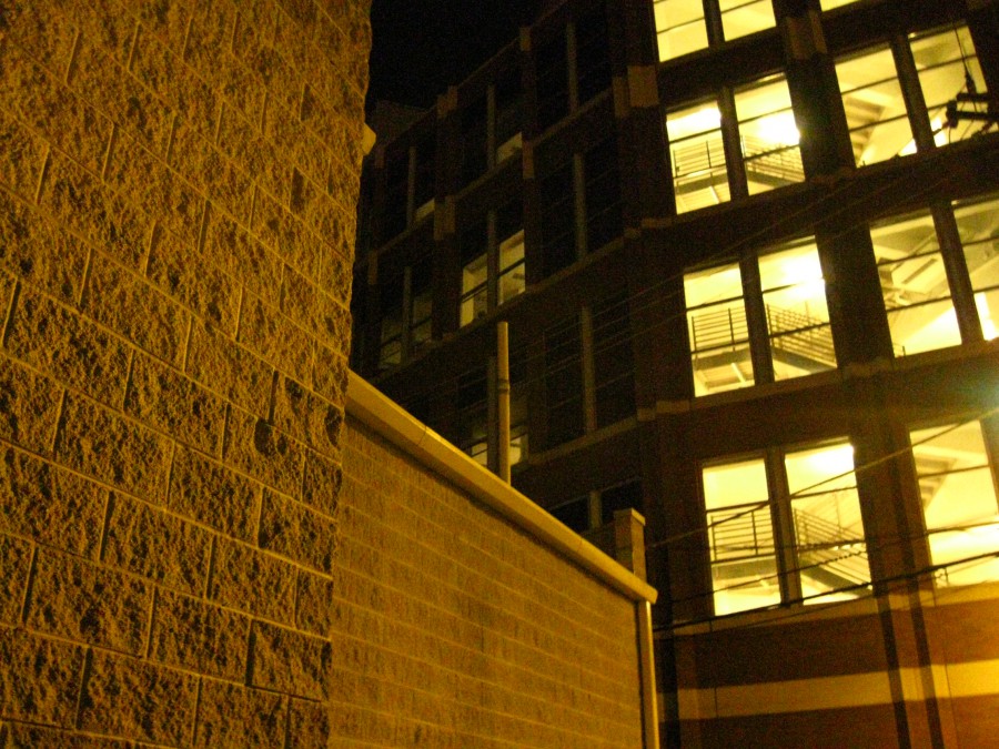 A shot of an architectural scene at night, showing textured concrete and brick walls in the foreground. Behind them, a tall modern building with bright yellow-lit windows reveals metal staircases on each floor. The contrast between the historic and modern elements, combined with the sharp lines and illumination, creates an engaging and slightly mysterious composition.