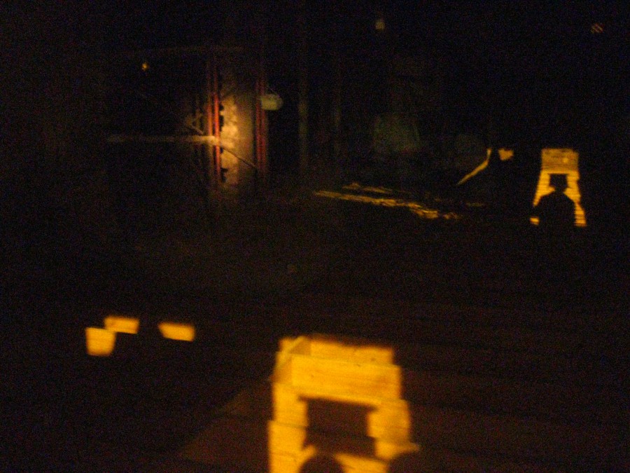 A dimly lit interior of a building captured at night through a glass window. The photo reveals a shadowy construction area with faint yellow light illuminating some elements. The left side shows wooden structures while shadows dominate the right, creating an eerie and mysterious vibe.