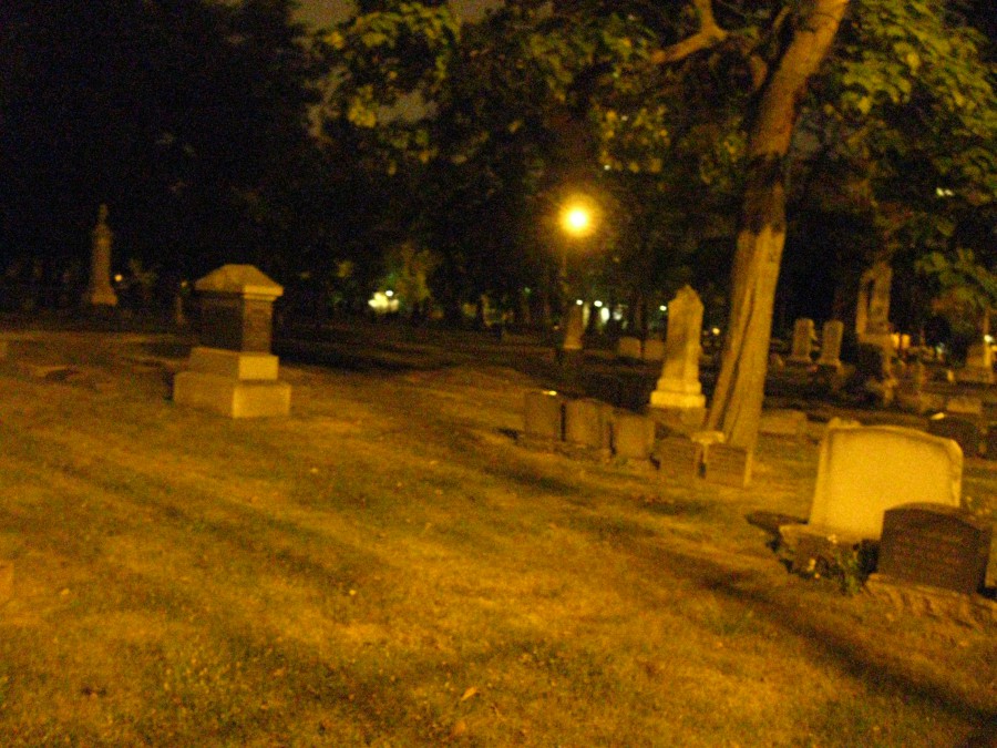 A nighttime graveyard scene featuring a grassy area with scattered tombstones and a large tree. A streetlight in the background casts an orange glow, partially illuminating the tombstones and creating long shadows that add to the spooky atmosphere. The foliage of the tree is thick and slightly blurred.