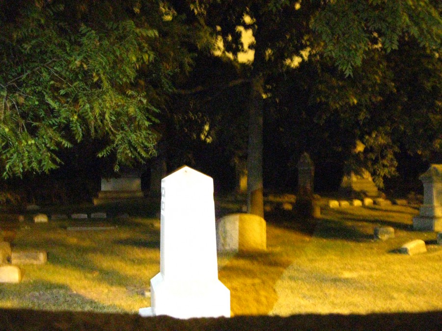 A flash-lit cemetery scene highlighting a single white tombstone monument that is brightly illuminated, contrasting with the surrounding darker tombstones and trees. The stark lighting gives the monument an almost spectral quality, with tree branches framing the background.