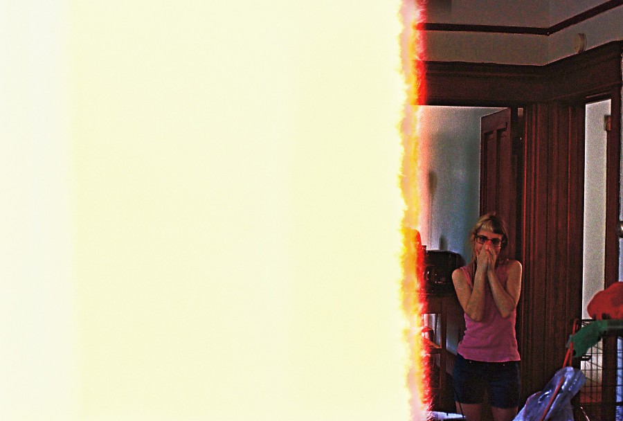 A striking image with most of the frame overtaken by an end-of-the-roll light leak in vivid shades of white, yellow, and orange. On the far right, Lauren is perfectly framed by the bright colors. She stands in a casual yet alluring pose, wearing a pink tank top and blue shorts. Her split-dyed hair is as vibrant as ever, and her hands are held over her face in a playful or surprised gesture. The background features an old-fashioned wood-paneled home interior, complete with visible doorframes and classic warm-toned trim, suggesting a vintage Chicago house.