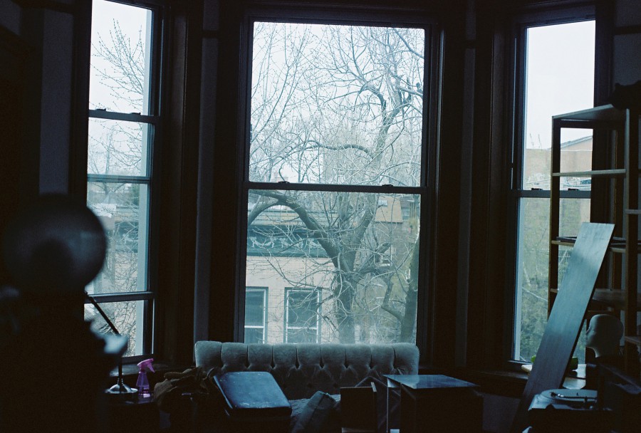 A view from inside a second-story Ukrainian Village apartment through large windows framed with dark wood. Outside, leafless trees and the muted façade of another apartment building suggest a chilly or early spring day. Inside, the clutter of moving is evident, with a tufted couch partially visible, a foam mannequin head on the right, and various packing boxes scattered throughout the room. A globe-like lamp on the left adds a touch of charm to the otherwise chaotic scene.