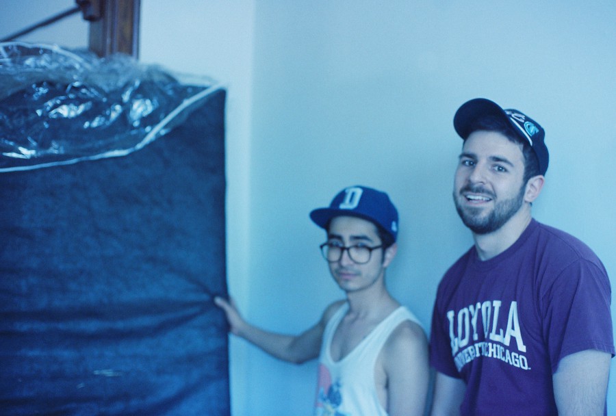 Alex, wearing glasses and a tank top, stands alongside Stanislav, who sports a Loyola University Chicago t-shirt. Both are in mid-action as they help move furniture. Alex is holding up a large, padded piece of furniture wrapped in protective material. The bright, diffused lighting creates soft shadows, and the background shows plain walls with hints of architectural wood trim typical of Chicago homes.