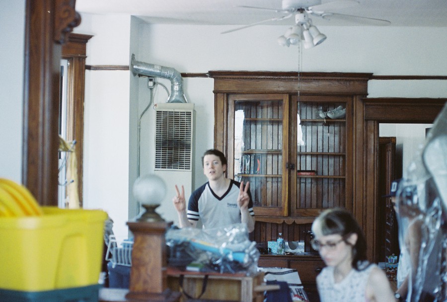 A lively and chaotic scene inside the house, where Brandon flashes two peace signs from the back of the room, wearing a casual shirt and grinning. Lauren is visible closer to the camera but slightly blurred, her split-dyed hair still discernible. The space is filled with plastic totes, wooden built-ins, and various household items scattered about. A vent and pipe on the wall add to the raw and lived-in feel of the room.