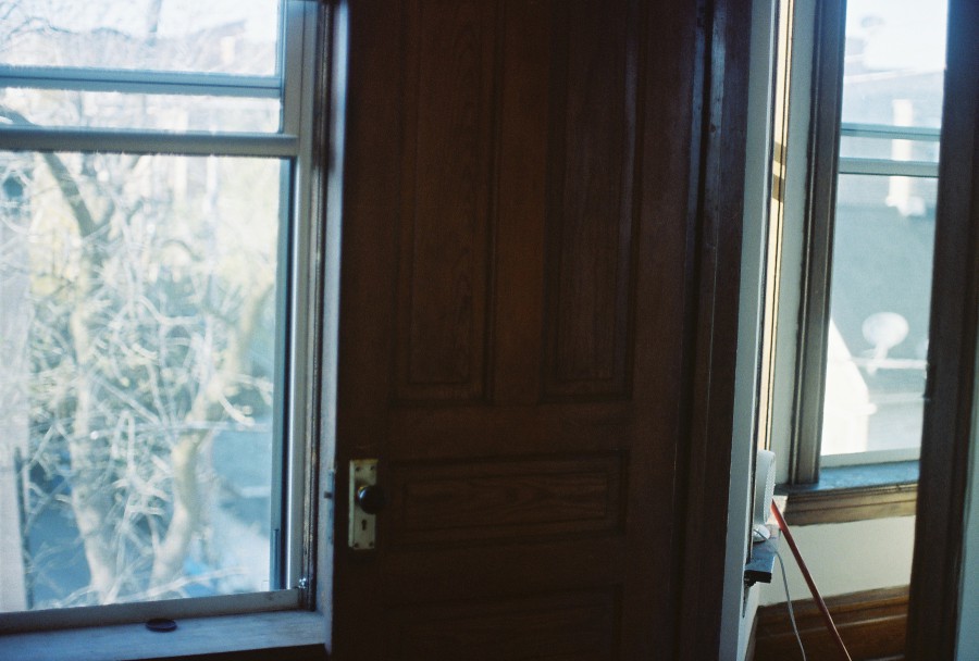 A tricky shot featuring a partially open door leading out from a bedroom. The doorframe on the right divides the image, with the left side revealing the interior of the room and a window that looks out onto the neighborhood and trees. The soft blue lighting and classic wood details evoke a sense of calm within the slight disarray of the moving process.