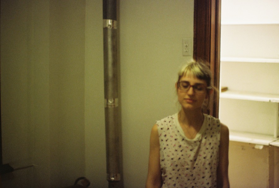 Lauren stands near an empty white pantry in a softly lit room, her split-dyed hair in shades of blonde and dark brown framing her face. She wears glasses and a sleeveless white shirt with a small dotted pattern, her eyes closed in a serene expression. Behind her, a metal pipe runs vertically along the wall.