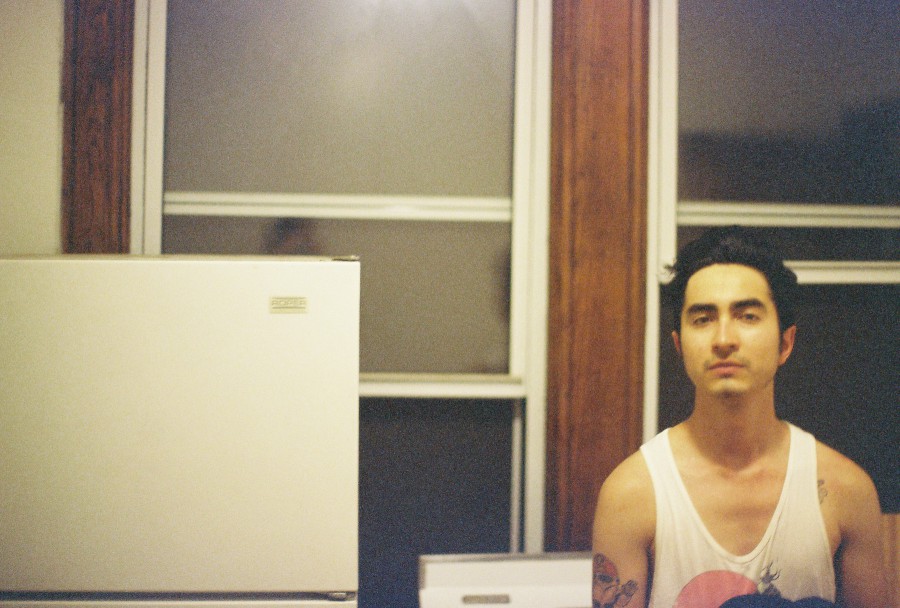 A kitchen scene, with a refrigerator prominently in the left foreground. Next to it stands Alex, wearing his white graphic tank top. The background features large windows framed by warm wood trim, allowing faint outdoor light to filter in. The scene is casual and unposed, capturing a candid moment of the moving night.
