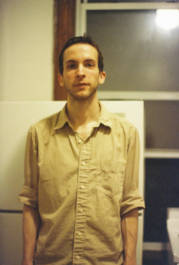 Ethan (me!), captured in a warmly lit kitchen with a slightly grainy texture that adds to the film-like aesthetic. I stand in front of a white refrigerator, dressed in a tan button-up shirt with rolled-up sleeves. The soft indoor lighting casts a gentle glow on my face. Behind me, large windows with dark trim show the reflection of the indoor light against the dark nighttime windows.