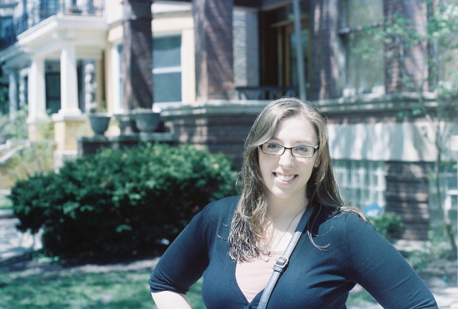 Outdoors in Rogers Park, Rebecca smiles brightly in the sunlight. She has long, light brown hair, slightly damp, and wears glasses with a confident expression. Her black cardigan and coral top complement the greenery and brick architecture behind her, the mid-day light casting soft shadows on the textured surroundings.