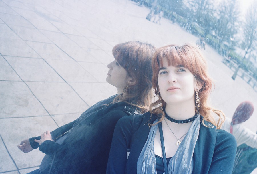 Erin leans against the reflective surface of Cloud Gate, also known as The Bean, in Millennium Park. Her vivid red hair falls to her shoulders, paired with striking dark brows and a spiky black choker. The reflection of her profile mirrors her pose against the sleek, polished metal.