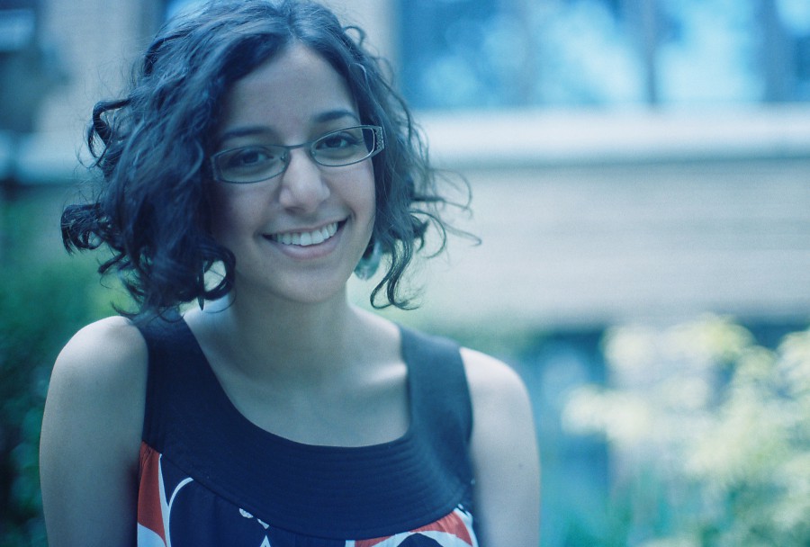 Annie's soft curls frame her face as she smiles in an outdoor shot. She wears glasses and a chic black top with colorful patterns, the bright hues echoing the soft greens and muted brick tones in the blurred background.