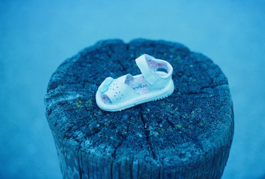 A tiny, white baby sandal with floral cutouts rests delicately atop a weathered wooden post. The pastel pink lining of the sandal contrasts with the rough texture of the wood. The background fades into a gentle blur of blue hues.