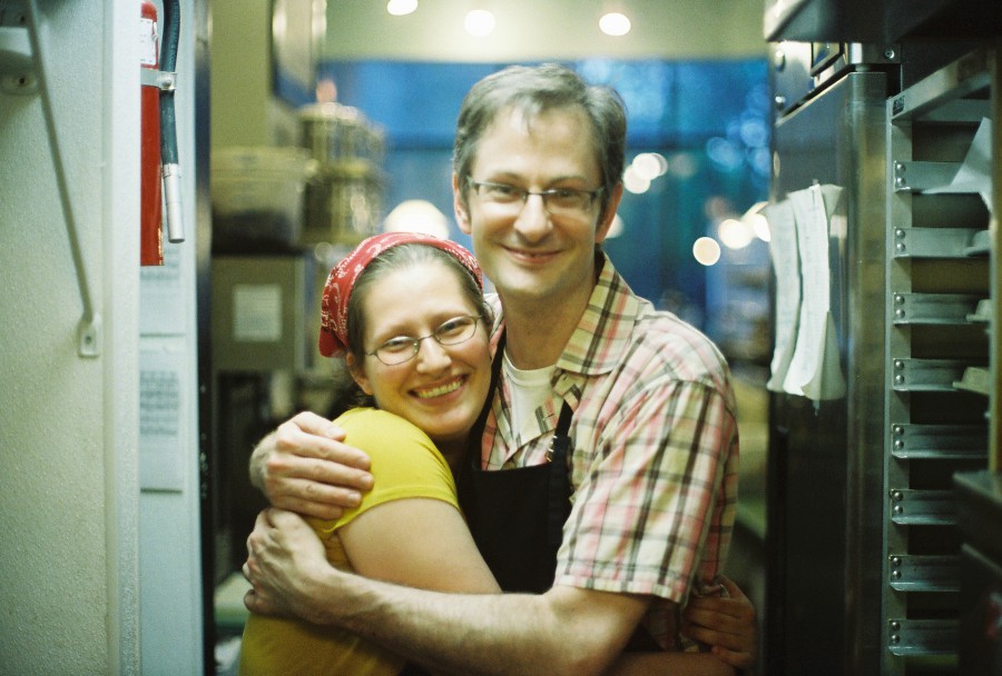 Victoria and Scott hug warmly in the back of a cozy coffee shop. Victoria, with a red bandana and glasses, beams with happiness, while Scott, in a checkered shirt and apron, smiles contentedly. The softly lit scene features coffee racks, pastry shelves, and warm wooden details, creating an inviting, bustling atmosphere.