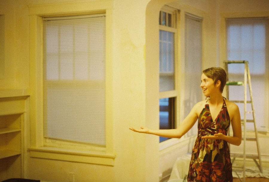In a freshly painted apartment in Rogers Park, Anna stands mock-presenting the space with outstretched hands. She wears a vibrant floral sundress, her short hair styled neatly. The walls are a white but appear pale yellow due to the 35mm film and lighting, and the room contains simple elements like blinds, a ladder, and a drop cloth on the floor, suggesting recent renovations.