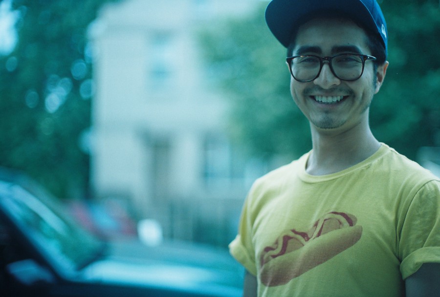 A 35mm film portrait shot with a vintage Petri SLR on the Fourth of July, 2013, showing a young man named Alex outdoors in West Town, Chicago. He’s smiling brightly with deep dimples, wearing black-rimmed glasses, a navy blue baseball cap, and a yellow T-shirt printed with a giant hotdog—perfectly kitschy Americana. The background is softly out of focus, showing a residential building and a parked car, all bathed in a teal-blue film tint. The bokeh effect and shallow depth of field make Alex pop warmly from the background, with a casual joy that captures the laid-back summer holiday vibe.