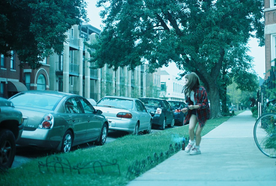 A dynamic street photo taken on 35mm film with a Petri SLR, showing Lauren in mid-movement on a West Town sidewalk just after lighting a bottle rocket. She’s turned slightly away, beginning to run as the firework smokes and hisses behind her, visible near her heels on the concrete. Lauren wears a red plaid shirt and sneakers, her long hair split-dyed—though here only the brunette side is visible from the camera angle. She looks beautiful, caught in motion, with a tree-lined street of cars and modern apartment buildings in the background. The color has a distinct cool cast from the film stock, adding a timeless and slightly dreamy atmosphere.