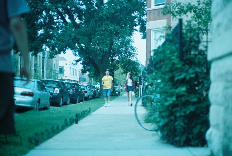 A 35mm film photo capturing Alex and Lauren walking back down the same Chicago sidewalk toward the photographer. Alex wears his yellow hotdog shirt and shorts, while Lauren—walking beside him—is effortlessly stunning in her plaid shirt and short shorts. She’s smiling slightly, framed by the trees overhead. In the blurred foreground at the far left, part of another person’s torso and arm are visible, giving the image a candid, slightly voyeuristic energy. A row of parked cars and multi-unit buildings line the quiet neighborhood block, giving the setting a chill, residential summer feel.