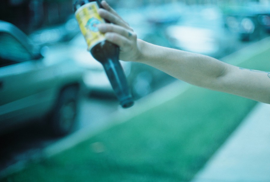 A creative close-up photo shot on 35mm film showing a single outstretched arm pouring out a glass bottle of Jarritos soda. Though the bottle appears to be pouring, it’s actually releasing smoke or ash from a recently lit firework—though the smoke itself is faint and not easily visible in the image. The background is blurred but shows grass and parked cars along a residential street. The person's skin is light-toned, and there’s a subtle tattoo on their arm. The soft focus and blue film cast give the image a dreamy, ambiguous feel—equal parts suburban mischief and filmic texture.