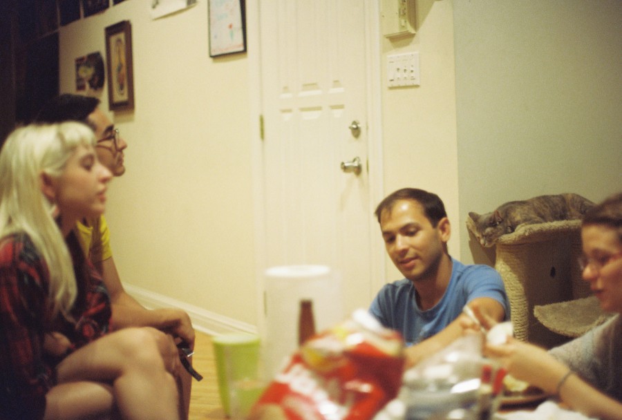 A warmly lit indoor snapshot taken on 35mm film showing a group of friends during a Fourth of July house hangout in West Town. From left to right: Lauren (visible with the blonde side of her split-dyed hair, sitting with her legs curled up, looking serene and beautiful), Alex (behind her, wearing the yellow hotdog shirt), Justyn (sitting cross-legged on the floor in a blue shirt, smiling slightly), Darcy the gray tabby cat lounging lazily on a carpeted cat tree in the background, and Amanda on the far right, mid-conversation and focused on making Jello shots. In the foreground, snack bags, bottles, and plastic cups cover the table, adding to the cozy, casual party vibe.
