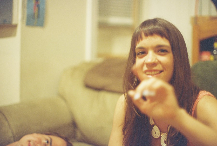 An indoor 35mm film photo focused on Caitlin, seated on a couch, smiling as she holds up a hand mid-gesture. She has long dark hair with bangs and wears a bold white necklace and a sleeveless coral top. The shot has a soft yellow warmth typical of indoor film lighting. To the lower left edge, only the top of Elliott’s head and part of his face are visible as he lies sideways out of focus, grinning playfully. Behind Caitlin, a beige couch and wall details—like a shelf and a small piece of hanging art—create a cozy, personal domestic backdrop.